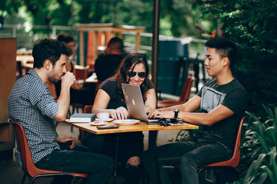 Three friends sitting on a shaded café terrace, around a laptop and cups.