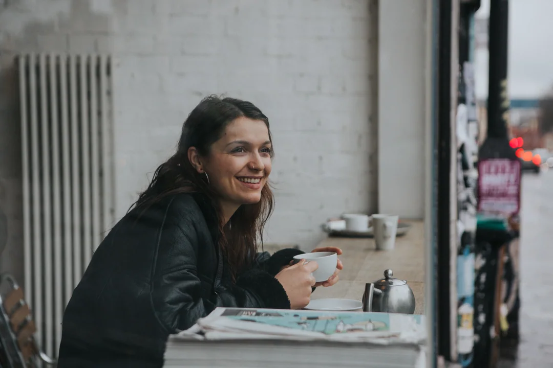 Une femme sourit, une tasse à la main, à la fenêtre d’un café.