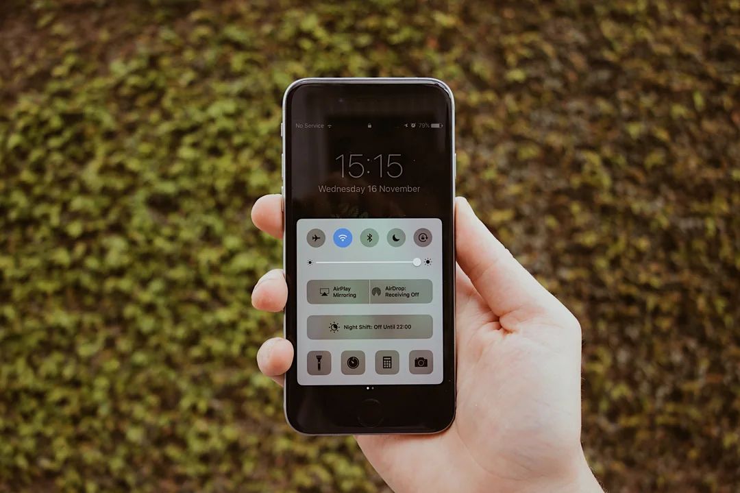 Une main tient un téléphone devant une haie de verdure.